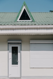 A building facade with a white door and a closed window shutter. Above these, there is a green tiled roof featuring a triangular dormer window with a black frame.