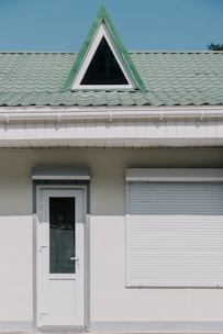 A building facade with a white door and a closed window shutter. Above these, there is a green tiled roof featuring a triangular dormer window with a black frame.