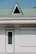 A building facade with a white door and a closed window shutter. Above these, there is a green tiled roof featuring a triangular dormer window with a black frame.