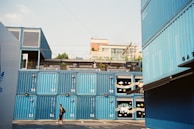Stacked blue shipping containers are arranged in an outdoor area with a man walking in front of them. The containers have numbers and some are decorated with colorful designs such as stars and rainbows. In the background, a building with balconies and some greenery on top can be seen under a clear sky.