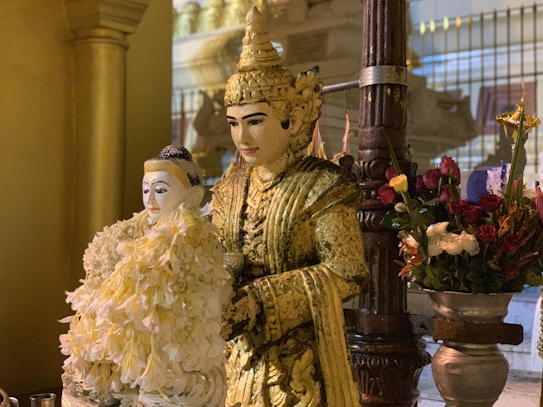 A golden statue adorned with intricate decorations, holding a smaller, white statue, surrounded by a display of vibrant flowers in metallic vases. The scene is illuminated with soft lighting, enhancing the ornate details of the statues and the rich colors of the flowers.