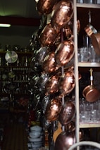 A variety of copper and metal pans are neatly hung on a store rack, alongside shelves filled with glassware and ceramic bowls. The setting appears to be a kitchenware shop with dim lighting, emphasizing the reflective surfaces of the pots and pans.