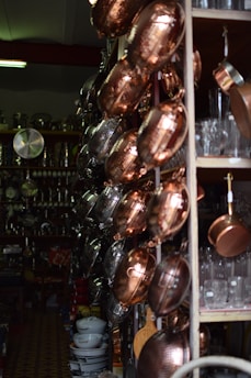 A variety of copper and metal pans are neatly hung on a store rack, alongside shelves filled with glassware and ceramic bowls. The setting appears to be a kitchenware shop with dim lighting, emphasizing the reflective surfaces of the pots and pans.
