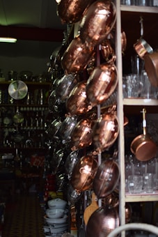 A variety of copper and metal pans are neatly hung on a store rack, alongside shelves filled with glassware and ceramic bowls. The setting appears to be a kitchenware shop with dim lighting, emphasizing the reflective surfaces of the pots and pans.