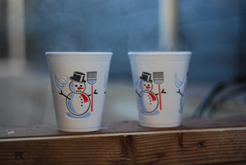 Two white styrofoam cups featuring a cheerful snowman design are positioned on a wooden surface. The snowmen have black top hats, red scarves, and are holding brooms. A hint of steam emanates from the cups, suggesting warmth within a cold setting.