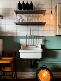 A vintage-style kitchen scene featuring a white ceramic sink with copper piping against a green and tiled wall. Above the sink, there are two shelves; the top shelf holds black bottles, while the bottom shelf has clear glass cups and white coffee mugs with a red logo. A warm Edison bulb hangs to the right, and a wooden chair is partially visible to the left. Below the sink, a round, decorative heater emits a warm orange glow.