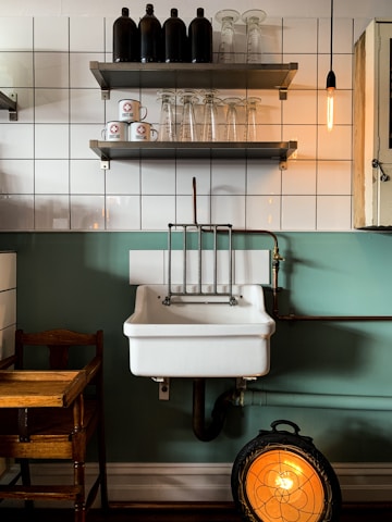 A vintage-style kitchen scene featuring a white ceramic sink with copper piping against a green and tiled wall. Above the sink, there are two shelves; the top shelf holds black bottles, while the bottom shelf has clear glass cups and white coffee mugs with a red logo. A warm Edison bulb hangs to the right, and a wooden chair is partially visible to the left. Below the sink, a round, decorative heater emits a warm orange glow.