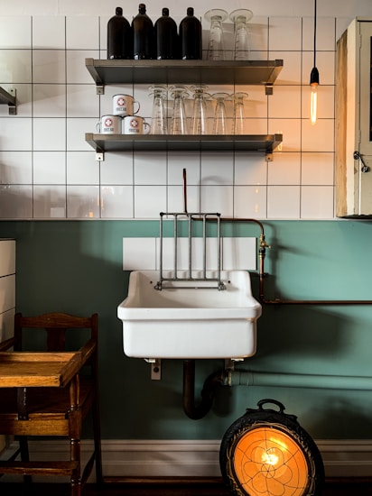 A vintage-style kitchen scene featuring a white ceramic sink with copper piping against a green and tiled wall. Above the sink, there are two shelves; the top shelf holds black bottles, while the bottom shelf has clear glass cups and white coffee mugs with a red logo. A warm Edison bulb hangs to the right, and a wooden chair is partially visible to the left. Below the sink, a round, decorative heater emits a warm orange glow.