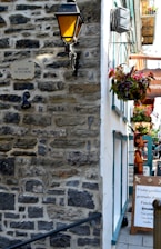 A narrow passageway with a rustic stone wall on the left, featuring an antique metal lantern. Below the lantern, a sign reads 'Escalier du Cul-de-Sac'. To the right, a building with white and teal details displays hanging flower baskets and signs advertising guided tours.