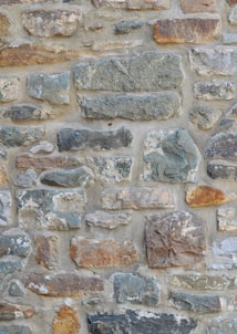 A textured stone wall composed of irregularly shaped stones with varying sizes and colors. The stones are organized in a random pattern and joined together with mortar. The surface displays a rugged look with earthy tones and subtle shadows, hinting at natural variation.