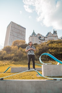 A person stands on a small concrete platform in an open grassy area with modern sculptures nearby. In the background, there are lush green trees and a tall high-rise building next to a historic stone building with pointed roofs under a partly cloudy sky.