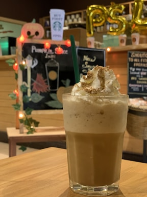 A glass filled with a frothy iced drink topped with whipped cream and cinnamon is prominently placed on a wooden table. In the background, a chalkboard displays a drink menu with festive decorations such as a carved pumpkin shape and orange lights. The interior setting features warm lighting and promotional signs, creating a cozy ambiance.