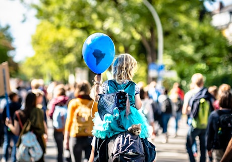 A young child with blonde hair sits on someone's shoulders, holding a blue balloon with a dark shape resembling a continent. The child wears a bright turquoise dress and has a small stuffed animal attached to a backpack. The scene takes place outdoors in a crowd of people, suggesting a participating march or protest. The blurred background is filled with trees and sunlight, creating a lively atmosphere.