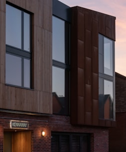 A modern building facade featuring large reflective windows and a mix of materials, including wood paneling and rust-colored metal cladding. A warmly lit entrance is visible, and there is a sign with the word 'KENNAWAY' above the door. The sky is clear with a subtle gradient of colors indicating a sunset or sunrise.