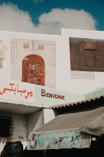 A wall displays a painted image of an archway with intricate decorations. Below the wall, a sign reads 'Bienvenue' alongside Arabic script. A muted blue and beige awning partially covers the view of a window and barrels at the bottom of the image.