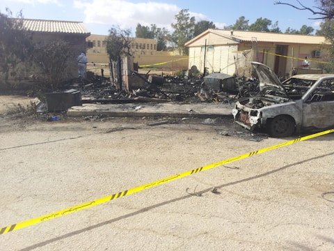 A burned-out vehicle sits on the side of a road surrounded by debris and charred remains of a structure. Yellow caution tape cordons off the area, indicating it is a restricted zone. In the background, two buildings are visible, with trees and a partly cloudy sky overhead. Some individuals wearing white protective suits are examining the scene.
