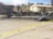 A burned-out vehicle sits on the side of a road surrounded by debris and charred remains of a structure. Yellow caution tape cordons off the area, indicating it is a restricted zone. In the background, two buildings are visible, with trees and a partly cloudy sky overhead. Some individuals wearing white protective suits are examining the scene.