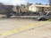 A burned-out vehicle sits on the side of a road surrounded by debris and charred remains of a structure. Yellow caution tape cordons off the area, indicating it is a restricted zone. In the background, two buildings are visible, with trees and a partly cloudy sky overhead. Some individuals wearing white protective suits are examining the scene.