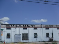 A large, industrial-style building with a white facade and several small windows. The words 'HOLLOWAY WIRE ROPE' are prominently displayed along the top in large, black letters. The building is bordered by a stop sign and power lines, with a bright blue sky and a few clouds in the background.