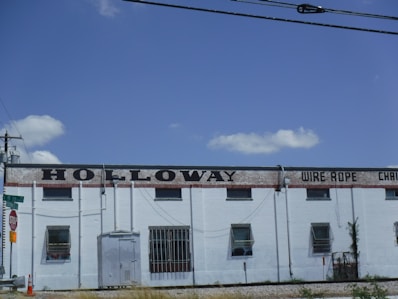 A large, industrial-style building with a white facade and several small windows. The words 'HOLLOWAY WIRE ROPE' are prominently displayed along the top in large, black letters. The building is bordered by a stop sign and power lines, with a bright blue sky and a few clouds in the background.