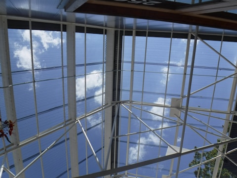 A perspective looking up at a glass or acrylic roof structure with a grid of metal beams framing sections of sky. The metal beams intersect at various angles, forming a geometric pattern against a backdrop of blue sky and scattered white clouds. A small portion of a leafy tree and some red flowers are visible at the edges.