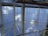 A perspective looking up at a glass or acrylic roof structure with a grid of metal beams framing sections of sky. The metal beams intersect at various angles, forming a geometric pattern against a backdrop of blue sky and scattered white clouds. A small portion of a leafy tree and some red flowers are visible at the edges.