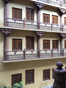 An architectural facade featuring multiple levels with wooden balconies and windows. The balconies are adorned with intricate geometric designs and each level is supported by thick pillars. The windows have a classic wooden design with lattice patterns. The building wall is painted in a light yellow color, and some greenery is visible at the base.