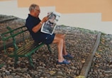 An elderly man is seated on a green bench in a cobblestone area, reading a magazine featuring a fashion advertisement. He wears a dark shirt and shorts, paired with blue sandals.
