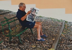 An elderly man is seated on a green bench in a cobblestone area, reading a magazine featuring a fashion advertisement. He wears a dark shirt and shorts, paired with blue sandals.