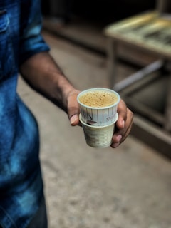 A person is holding a cup filled with coffee or tea. The person is wearing a denim jacket, and the cup is made of disposable material with some text and illustrations.