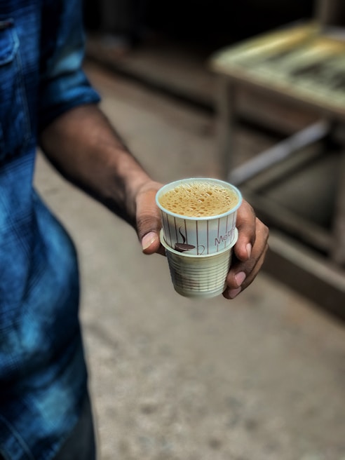 A person is holding a cup filled with coffee or tea. The person is wearing a denim jacket, and the cup is made of disposable material with some text and illustrations.