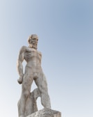 A stone statue of a bearded man with a muscular build standing in a powerful pose against a clear blue sky backdrop. The figure appears to be wearing no clothing and is depicted in a classical style.