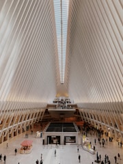A large, modern indoor space characterized by an impressive vaulted ceiling with rib-like structures stretching towards a skylight. The floor is bustling with people, suggesting a public or commercial area. There are small structures and booths, adding to the cosmopolitan atmosphere.