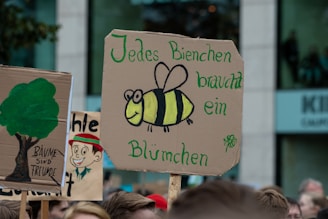 A crowd scene with various handmade signs held up, one prominently featuring a painted bee and a message in German about bees and flowers. Another sign shows a tree with a short slogan beneath it in German. The scene appears to be part of a protest or demonstration, likely for environmental or ecological causes.
