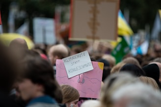 A large crowd of people gathers, holding various protest signs. In the foreground is a sign with German text, advocating for climate protection over coal pollution. The scene appears to be a protest or demonstration with colorful banners and signs visible in the background.