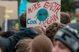 A crowd of people gathered at a protest. One person is holding a handmade sign featuring a cartoon Earth wearing sunglasses with the slogan 'Make Earth Cool Again' written in bold colors.
