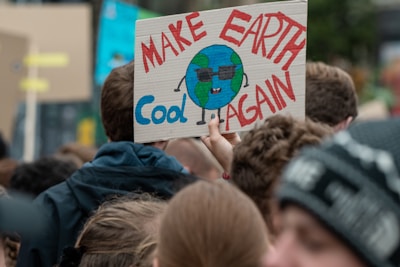 A crowd of people gathered at a protest. One person is holding a handmade sign featuring a cartoon Earth wearing sunglasses with the slogan 'Make Earth Cool Again' written in bold colors.