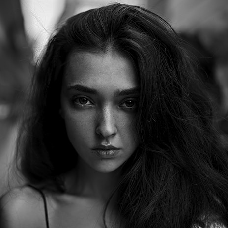 A black and white portrait of a woman with long, dark hair flowing freely around her face. Her expression is intense and serious, with deep, captivating eyes and slightly parted lips. The background is softly blurred, focusing attention on her facial features.