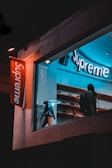 A storefront with a brightly lit sign displaying the brand name 'Supreme'. Inside, a person is looking at the display shelves filled with various items. There is a modern, stylish lamp on a stool near the window. The scene is captured during nighttime, giving it a cool, urban feel.