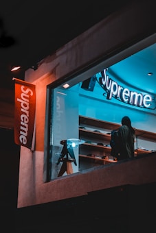 A storefront with a brightly lit sign displaying the brand name 'Supreme'. Inside, a person is looking at the display shelves filled with various items. There is a modern, stylish lamp on a stool near the window. The scene is captured during nighttime, giving it a cool, urban feel.