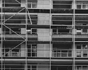 A black and white image of a building under construction featuring multiple levels of scaffolding and partially built balconies. The structure is predominantly concrete with visible metal framework for support.