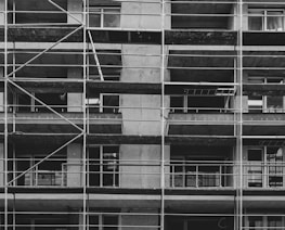 A black and white image of a building under construction featuring multiple levels of scaffolding and partially built balconies. The structure is predominantly concrete with visible metal framework for support.