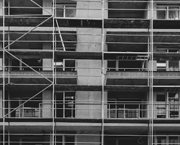 A black and white image of a building under construction featuring multiple levels of scaffolding and partially built balconies. The structure is predominantly concrete with visible metal framework for support.