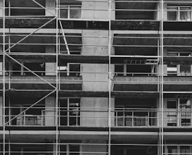A black and white image of a building under construction featuring multiple levels of scaffolding and partially built balconies. The structure is predominantly concrete with visible metal framework for support.