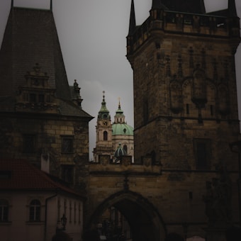 A view of historic architecture with tall, intricate towers and a distant dome topped with a cross. The buildings exhibit ornate stonework and structural details typical of medieval European design.
