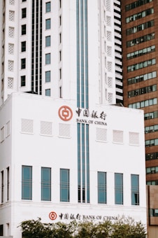 A tall, modern building with a white facade features the logo and name of the Bank of China. The structure includes rectangular windows arranged symmetrically and is flanked by another building with a darker, brown exterior. The architecture reflects a blend of contemporary and traditional Chinese elements.