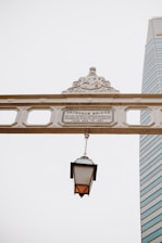 An ornate bridge sign with detailed embellishments and a historical plaque bears the name of 'Cavenagh Bridge'. A classic, lantern-style streetlamp hangs from the sign, providing a nostalgic element. In the background, a modern skyscraper with glass windows stands tall against a clear, overcast sky.