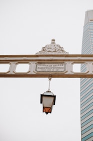 An ornate bridge sign with detailed embellishments and a historical plaque bears the name of 'Cavenagh Bridge'. A classic, lantern-style streetlamp hangs from the sign, providing a nostalgic element. In the background, a modern skyscraper with glass windows stands tall against a clear, overcast sky.
