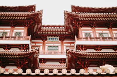 A traditional East Asian architectural structure with multiple levels, featuring ornate, upturned eaves and red and white detailing. The building includes intricate woodwork and a decorative plaque with Chinese characters.