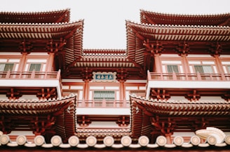 A traditional East Asian architectural structure with multiple levels, featuring ornate, upturned eaves and red and white detailing. The building includes intricate woodwork and a decorative plaque with Chinese characters.